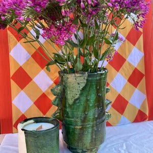 Puede incluir: Un jarrón cilíndrico alto de cerámica esmaltada en verde oscuro con crestas laterales prominentes, lleno de un gran ramo de flores moradas vibrantes, junto a un recipiente de vela de cerámica más pequeño a juego en verde y blanco con mecha de madera.