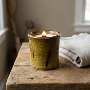 Op de afbeelding: Een brandende kaars in een cilindervormige, gespikkelde olijfgroene keramische houder met een getextureerd, onregelmatig oppervlak. De kaars heeft een enkele houten lont die een kleine vlam geeft, geplaatst in een dikwandige pottenbak.
