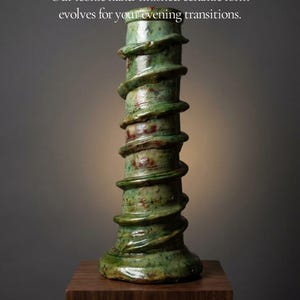 Op de afbeelding: Een hoge, cilindrische groene keramische vaas met een verhoogde spiraalstructuur die rond de buitenkant wikkelt. De tekst A NEW LIGHT ON OUR SIGNATURE SPIRAL en Our iconic hand-finished ceramic form evolves for your evening transitions. is zichtbaar.