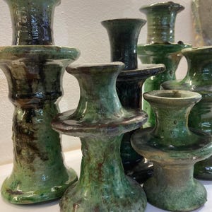Può includere: Una collezione di portacandele in ceramica fatti a mano in varie dimensioni e tonalità di verde. I portacandele hanno una finitura lucida e un aspetto unico e strutturato. Il design presenta una base svasata e una parte superiore ampia e piatta.
