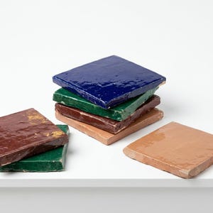 May include: A collection of square ceramic tiles with a glossy finish, featuring a stack of four tiles in deep blue, green, dark brown, and light tan, alongside two additional tiles in dark brown and green resting separately on the surface.