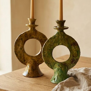 May include: Two ceramic candle holders with a circular, ring-shaped design and a textured, glossy finish. One holder is a muted olive green, while the other is a deeper, brownish-yellow tone. Each holds a single, slender, light beige taper candle.