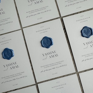 Op de afbeelding: Een verzameling witte kaarten met elegante kalligrafie en blauwe waszegels. De kaarten tonen de namen "YASSINE and AMAL" en de datum 14.11.2025, met extra tekst in een vreemde taal. De zegels hebben een bloemenontwerp.