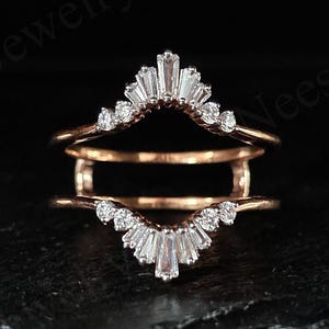 Puede incluir: Un protector de anillo de oro rosa con dos bandas curvas. Cada banda está adornada con pequeñas piedras blancas redondas y baguette. El protector de anillo está diseñado para acompañar un anillo de compromiso.