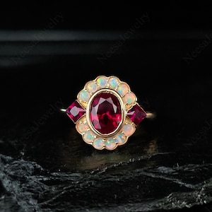 May include: A gold ring featuring a large oval ruby center stone, surrounded by a halo of opals and two smaller ruby accent stones. The ring has a vintage or antique style.
