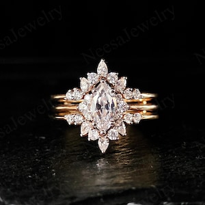 May include: A marquise-cut diamond engagement ring with a halo of smaller diamonds, set on a gold band. The ring is part of a three-piece set, with two additional gold bands. The jewelry is displayed against a dark background.