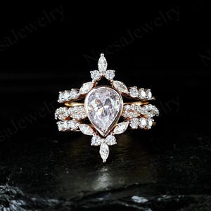 May include: A three-piece rose gold ring set. The engagement ring features a pear-shaped diamond center stone, surrounded by smaller diamonds and marquise-cut stones. The bands are adorned with pave-set diamonds. The rings are set against a black background.
