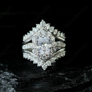 May include: A three-piece silver ring set with a large oval-cut center stone and numerous smaller stones. The ring is set against a black background, highlighting the sparkle of the stones. The set includes a band and two additional pieces.