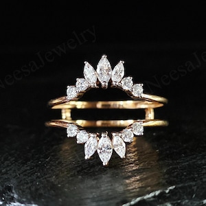 May include: A gold-coloured ring guard with a double band design. The top and bottom arches are decorated with clear, sparkling gemstones, including marquise and round cuts. The ring is set against a dark background.