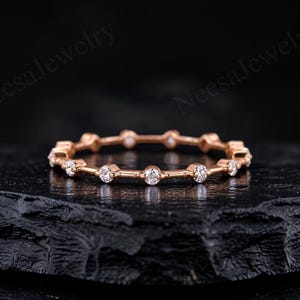 May include: A rose gold band ring featuring a series of evenly spaced, round, clear, faceted stones set in individual metal bezels. The ring has a thin, delicate profile with a polished finish, showcasing a repeating pattern of metal bars and stones.