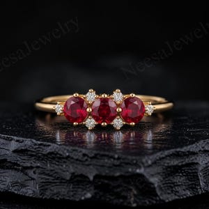 Pode incluir: Um anel de ouro com três rubis vermelhos de corte redondo dispostos horizontalmente no centro, acentuado por quatro pequenos diamantes brancos de corte redondo colocados entre e nas laterais dos rubis. O texto NeesaJewelry é visível ao fundo.