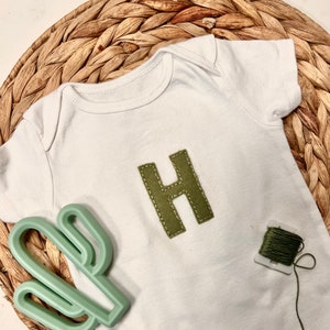 May include: A white baby onesie with a green felt letter H sewn on the front. A green cactus-shaped teether and a spool of green thread are also in the image.