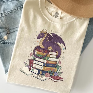 May include: Cream-coloured t-shirt with a purple dragon graphic sitting on books. The design includes roses, glasses, and open books. Light blue jeans and a tan hat are also visible.