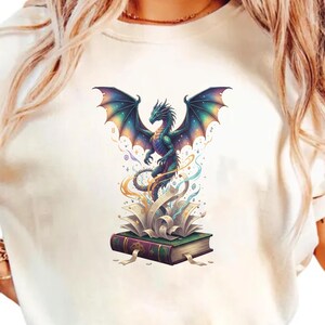 May include: Cream-coloured t-shirt with a colourful dragon illustration. The dragon has outstretched wings and sits atop an open book with swirling details. The dragon is mainly teal and purple.