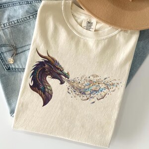 May include: Cream-colored t-shirt featuring a detailed dragon graphic in shades of purple, green, and gold. The dragon is exhaling a cloud of intricate designs. The shirt is paired with light-wash jeans, a necklace, and a tan hat.