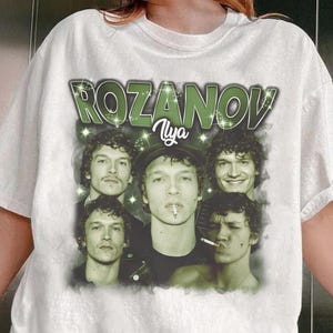 May include: White t-shirt featuring a graphic design with the text "ROZANOV" in green, along with the name "Ilya". The design includes multiple images of a man with curly hair. The shirt is a casual wear item.