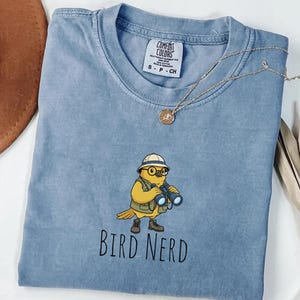 May include: A pale blue t-shirt featuring a cartoon bird wearing a hat and glasses, holding binoculars. The text "BIRD NERD" is printed below the bird. A gold necklace with a pendant is draped across the shirt. The shirt is a Comfort Colors brand.