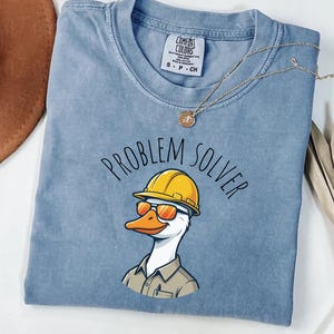 May include: A pale blue t-shirt featuring the text "PROBLEM SOLVER" and a cartoon duck wearing a yellow hard hat and sunglasses. The shirt has a crew neck and short sleeves. A gold necklace is draped across the shirt.