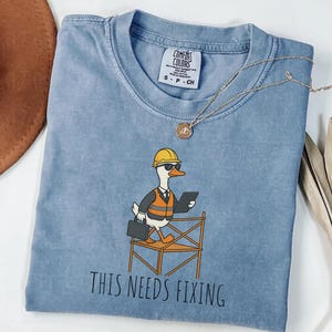 Op de afbeelding: Lichtblauw Comfort Colors T-shirt met een cartoon eend met een helm, zonnebril en een oranje vest, met een tablet en aktetas. De tekst "THIS NEEDS FIXING" staat eronder. Een gouden ketting ligt over het shirt.