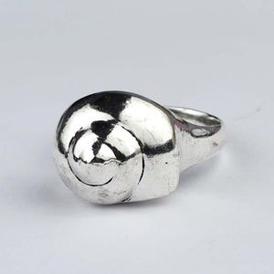 May include: A silver ring with a spiral design. The ring has a rounded, shell-like shape with a detailed spiral pattern on the face. The band is smooth and simple, and the ring is set against a white background.