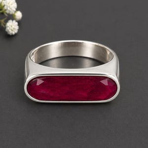 May include: A silver-toned ring featuring a horizontal, elongated oval setting. The centre of the ring contains a faceted, deep red gemstone with a textured surface. The band is smooth and polished, providing a clean, metallic finish to the piece.