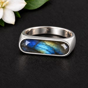 Puede incluir: Un anillo de plata con una piedra preciosa de labradorita facetada, horizontal y alargada, con destellos iridiscentes azules y dorados. La piedra está engastada en una banda de metal pulido y liso, junto a una flor blanca con hojas verdes.