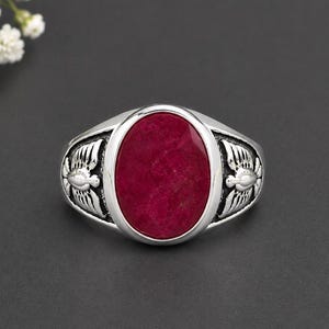 Könnte beinhalten: Ein silberner Ring mit einem großen, ovalen, tiefroten Stein in der Mitte. Die Ringschiene ist auf beiden Seiten mit detaillierten Metallarbeiten verziert, die stilisierte Flügel oder Federn darstellen, die von der Fassung ausgehen.