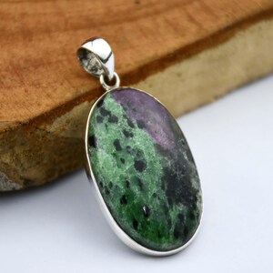 Può includere: Ciondolo ovale con una pietra lucida multicolore. La pietra presenta sfumature verdi, nere e viola, incastonata in una montatura in metallo color argento. Il ciondolo ha un anello nella parte superiore per una catena.