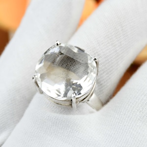 May include: A silver ring with a large, clear, faceted gemstone. The gemstone is cushion-cut and set in a simple silver band. The ring is shown against a white fabric background, emphasising its clarity and sparkle.