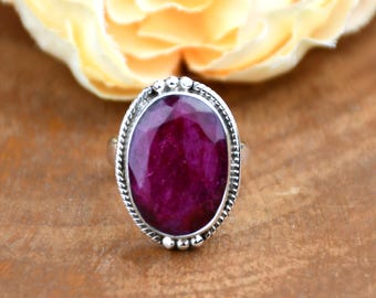 Puede incluir: Un anillo de plata con una gran piedra preciosa roja oscura de corte ovalado. La piedra preciosa está engastada en una banda de plata detallada con un diseño texturizado. El anillo se muestra sobre una superficie de madera, con una flor amarilla borrosa en el fondo.