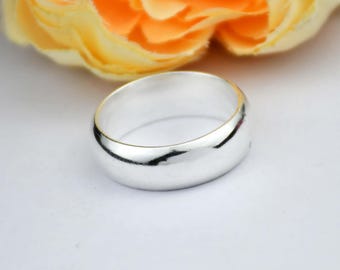 May include: A shiny, silver ring with a smooth, rounded design. The ring is set against a soft white background, with a blurred yellow and orange flower in the background. The ring's polished surface reflects light.