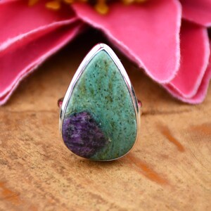 May include: A silver ring with a large, teardrop-shaped stone. The stone is mottled, with green and purple hues. The ring is set against a wooden surface, with a pink flower in the background.