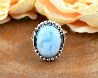 May include: A sterling silver ring featuring a large oval-shaped, light blue gemstone. The gemstone has a marbled appearance with shades of white and blue. The ring has a beaded border and is set against a blurred floral background.
