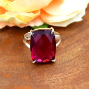 Puede incluir: Un anillo de plata con una gema grande, rectangular, facetada y de color rojo intenso. El anillo tiene una banda dorada debajo de la gema. El fondo está borroso, mostrando una flor y una superficie de madera.