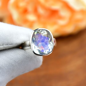 May include: A silver ring featuring a large, faceted, light-colored gemstone with iridescent blue and purple hues. The gemstone is set in a simple, polished silver band. The ring is held against a white fabric background.