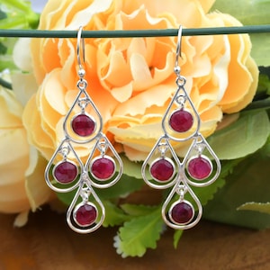 May include: Silver chandelier earrings with teardrop designs. Each earring features three tiers of ruby-red gemstones set within openwork frames. The earrings hang from a simple hook, showcasing a classic and elegant style.