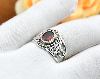 Puede incluir: Un anillo de plata con una piedra preciosa ovalada de color rojo intenso. El anillo presenta un diseño de filigrana detallado con pequeños detalles de cuentas alrededor de la piedra preciosa. El anillo se muestra sobre una superficie blanca.
