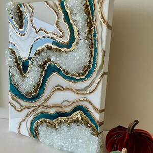 May include: An abstract canvas art piece with a geode design. The artwork features layers of white, teal, gold, and clear crystal accents. Two velvet pumpkins, one red and one green, are in the foreground.