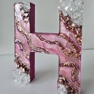 May include: A decorative letter "H" in a pink and purple color scheme, adorned with gold glitter and clear crystal accents. The letter has a textured, layered design, creating a unique, artistic appearance.