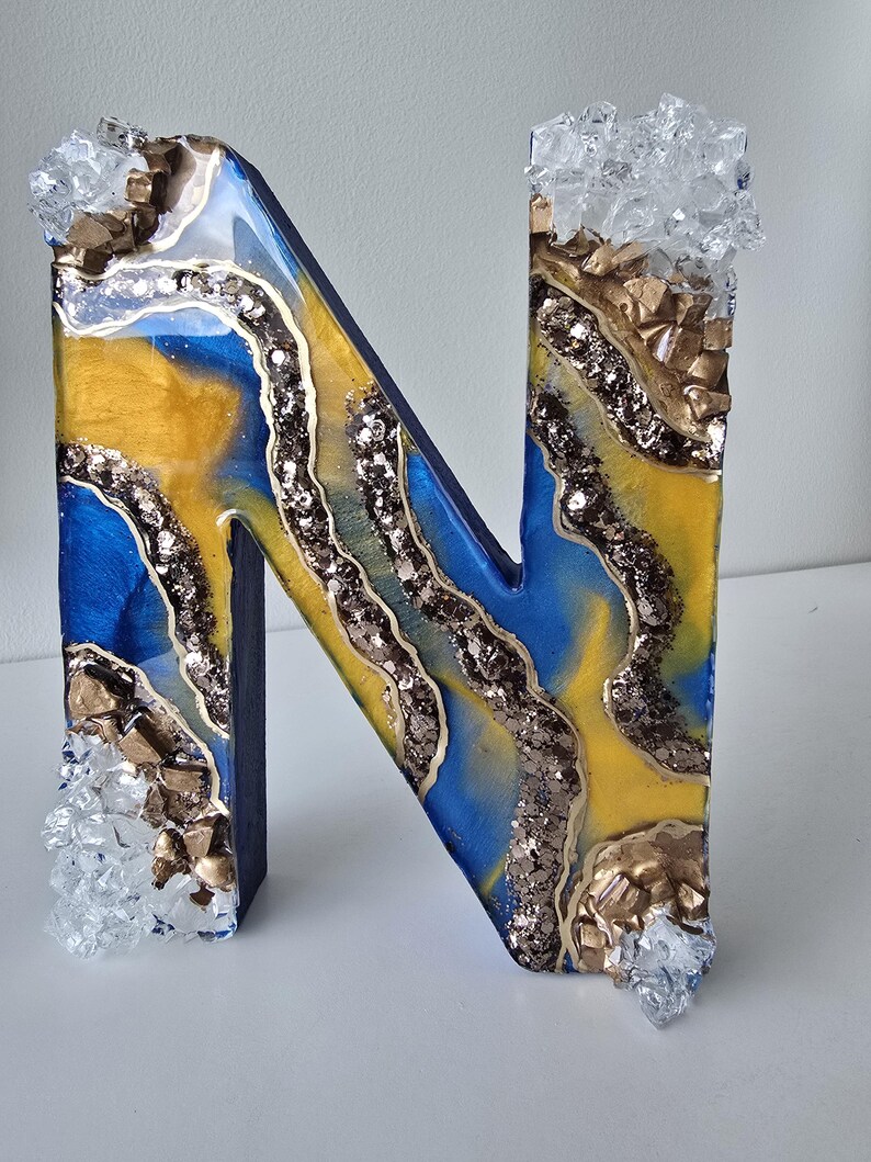 Puede incluir: Letra decorativa "N" con un dise&ntilde;o de resina azul, amarilla y dorada. La letra est&aacute; adornada con purpurina y detalles de cristal. La letra es un objeto decorativo.