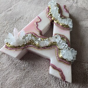 May include: A decorative letter "A" in a light pink and white color scheme, adorned with gold and pink glitter accents. Clear crystal clusters are embedded throughout the design, creating a unique, eye-catching aesthetic.
