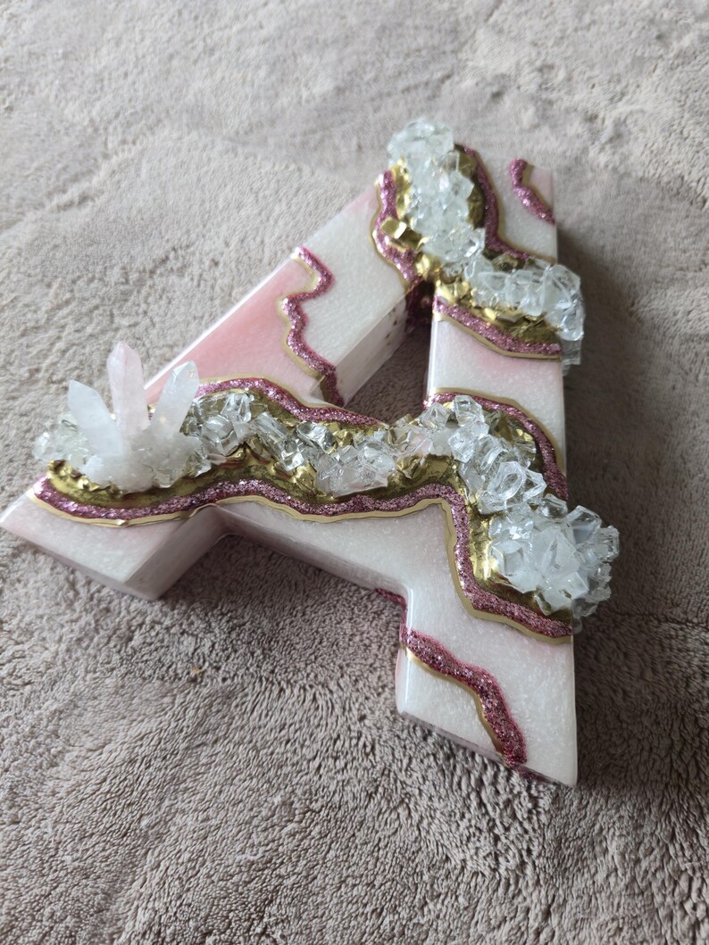 May include: A decorative letter "A" in a light pink and white color scheme, adorned with gold and pink glitter accents. Clear crystal clusters are embedded throughout the design, creating a unique, eye-catching aesthetic.