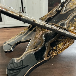 May include: A decorative, handcrafted stand with a black, gold, and silver resin design. The stand features a layered effect with gold leaf accents and a glossy finish. The edges are outlined in gold, creating a luxurious aesthetic.