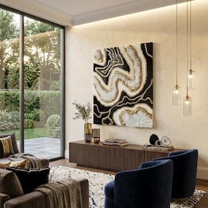 May include: A contemporary living room with a large abstract artwork in black, white, and gold. The room includes a dark wood console, two navy armchairs, and a patterned rug. Three pendant lights hang from the ceiling, illuminating the space.
