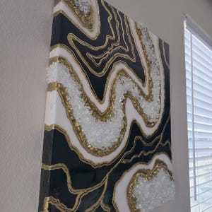 May include: Abstract wall art with a geode design. The artwork features a black and white pattern with gold glitter accents. Clear, crushed glass is embedded along the edges, adding texture. The canvas is mounted on a wooden frame, creating a striking visual.