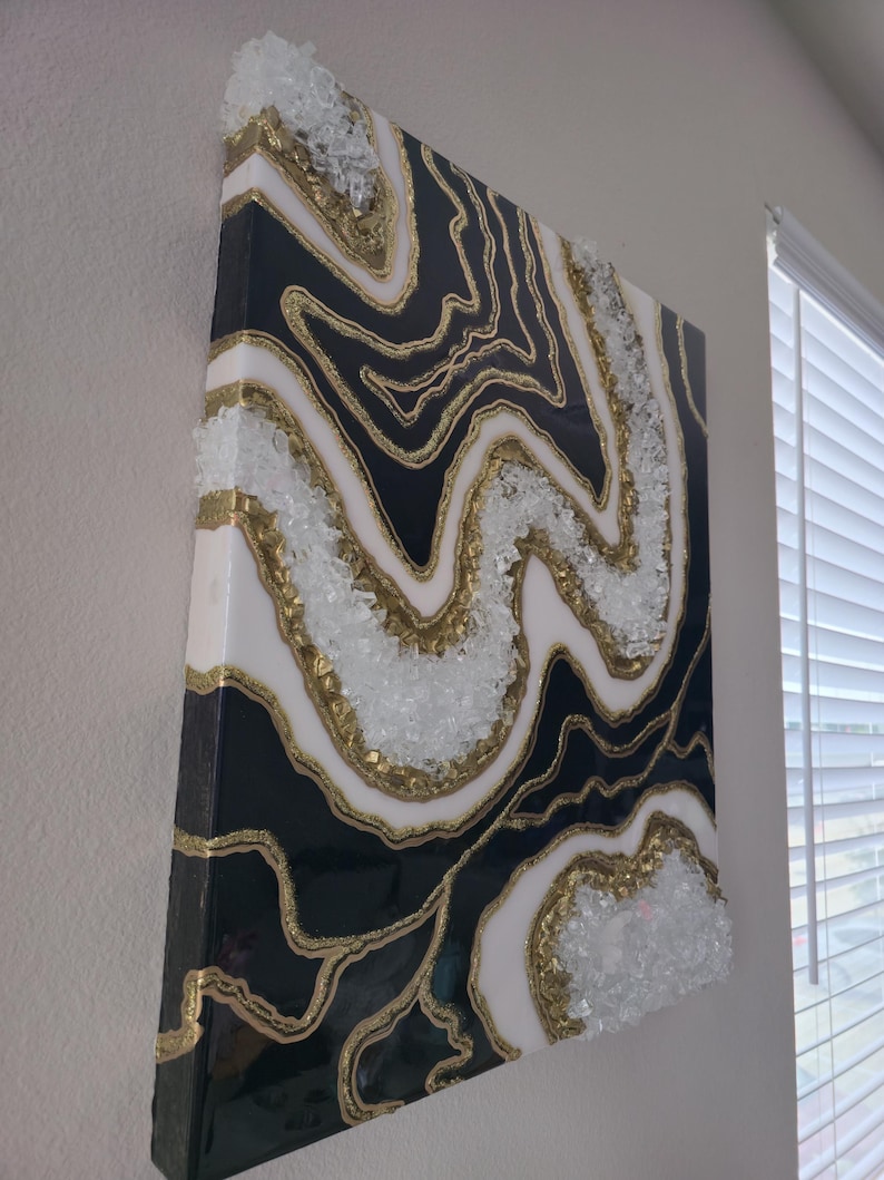 May include: Abstract wall art with a geode design. The artwork features a black and white pattern with gold glitter accents. Clear, crushed glass is embedded along the edges, adding texture. The canvas is mounted on a wooden frame, creating a striking visual.