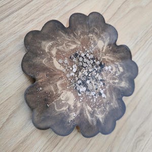 K&ouml;nnte beinhalten: Ein bl&uuml;tenf&ouml;rmiger Untersetzer mit einem dunkelbraunen und goldenen Harzdesign. Der Untersetzer hat einen gewellten Rand und ist mit silbernen Glitzerakzenten in der Mitte verziert. Der Untersetzer liegt auf einer Holzoberfl&auml;che.