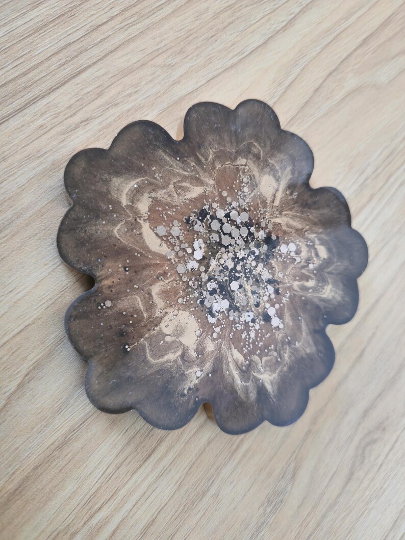K&ouml;nnte beinhalten: Ein bl&uuml;tenf&ouml;rmiger Untersetzer mit einem dunkelbraunen und goldenen Harzdesign. Der Untersetzer hat einen gewellten Rand und ist mit silbernen Glitzerakzenten in der Mitte verziert. Der Untersetzer liegt auf einer Holzoberfl&auml;che.