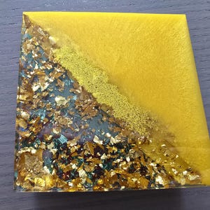 Pode incluir: Base para copos quadrada em resina com um design vibrante. A base apresenta uma divis&atilde;o diagonal: um lado &eacute; amarelo s&oacute;lido, o outro &eacute; uma mistura de folha de ouro e verde-azulado. Uma linha dourada texturizada separa as duas se&ccedil;&otilde;es.