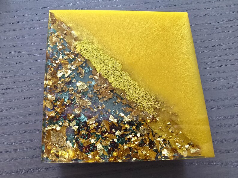 Pode incluir: Base para copos quadrada em resina com um design vibrante. A base apresenta uma divis&atilde;o diagonal: um lado &eacute; amarelo s&oacute;lido, o outro &eacute; uma mistura de folha de ouro e verde-azulado. Uma linha dourada texturizada separa as duas se&ccedil;&otilde;es.